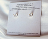 Pair of gold earrings on a card with text about hypoallergenic and tarnish-resistant material, displayed on a white fabric background.
