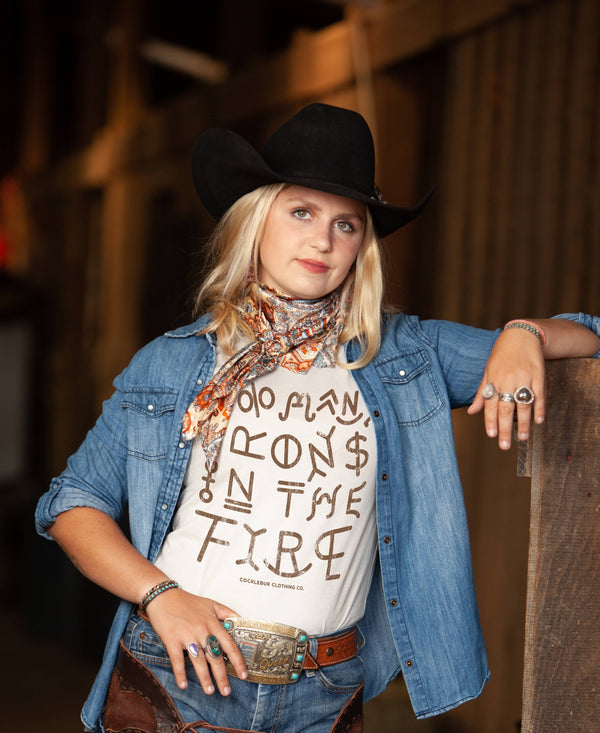 A person wearing a graphic tee with the text TOO MANY IRONS IN THE FIRE' printed on it, paired with a denim jacket and a cowboy hat.