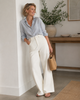 Woman in light blue shirt and white barrel jeans standing indoors with a plant and wooden table in the background.