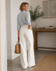 Woman in light blue shirt and white barrel jeans holding a brown bag in a room with a wooden table and plant.