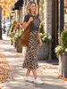 A woman stands on a city sidewalk wearing a dark gray graphic t-shirt, a leopard-print midi skirt, white sneakers, and carrying a large brown fringe shoulder bag.
