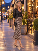 A woman stands on a city sidewalk at dusk wearing a black leather jacket, black satin top, leopard-print midi skirt, and black strappy heels, holding a small black clutch.