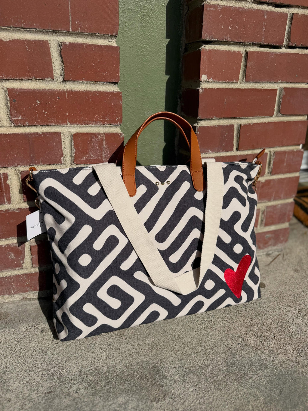 A canvas tote bag with a black and off-white geometric maze pattern, brown leather handles, and a red heart patch on the side, photographed outdoors against a brick wall.