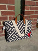 A canvas tote bag with a black and off-white geometric maze pattern, brown leather handles, and a red heart patch on the side, photographed outdoors against a brick wall.