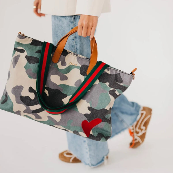 A person carrying a large camouflage tote bag with green and red web-style handles, light brown leather handles, and a red heart detail on the front, against a clean white background.