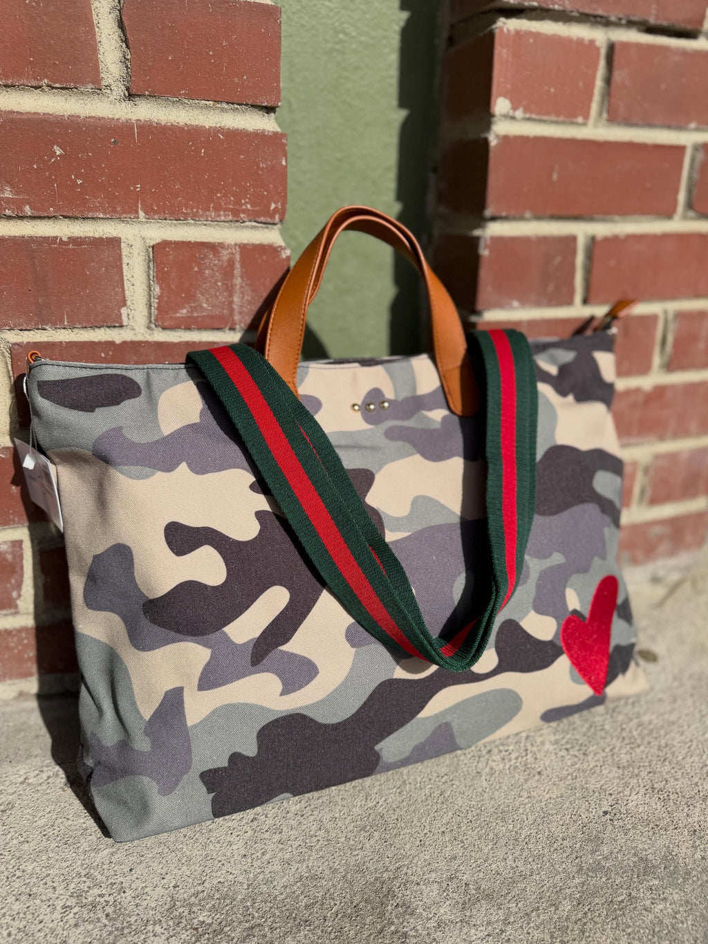 A canvas tote bag with a gray, beige, and black camouflage pattern, featuring green and red web-style shoulder straps, brown leather handles, and a red heart patch on the bottom right, photographed outdoors against a brick wall.