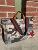 A canvas tote bag with a gray, beige, and black camouflage pattern, featuring green and red web-style shoulder straps, brown leather handles, and a red heart patch on the bottom right, photographed outdoors against a brick wall.