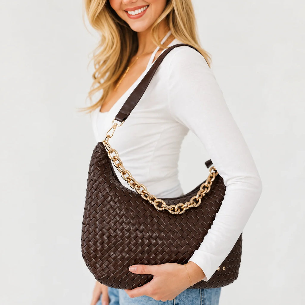 A woman wearing a white long-sleeve top and blue jeans carries a dark brown woven leather shoulder bag with a chunky gold chain strap against a plain white background.