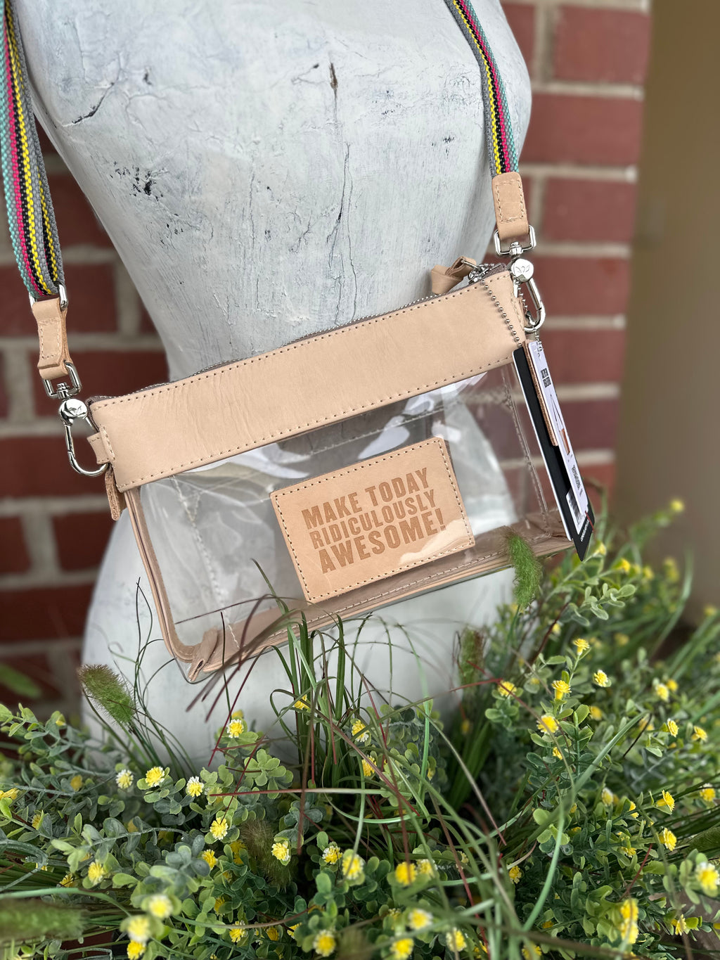 A clear plastic crossbody bag with a genuine leather trim and strap, displayed on a white mannequin torso with green and yellow flowers in the foreground and a brick wall behind it. The bag features a clear front pocket with a beige patch that reads 'MAKE TODAY RIDICULOUSLY AWESOME!' 