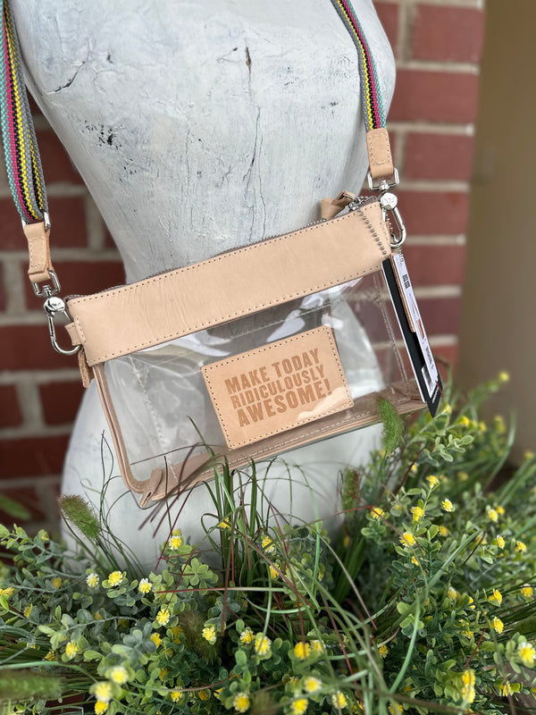 A clear plastic crossbody bag with a genuine leather trim and strap, displayed on a white mannequin torso with green and yellow flowers in the foreground and a brick wall behind it. The bag features a clear front pocket with a beige patch that reads 'MAKE TODAY RIDICULOUSLY AWESOME!' 