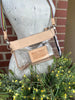 A clear plastic crossbody bag with a genuine leather trim and strap, displayed on a white mannequin torso with green and yellow flowers in the foreground and a brick wall behind it. The bag features a clear front pocket with a beige patch that reads 'MAKE TODAY RIDICULOUSLY AWESOME!' 