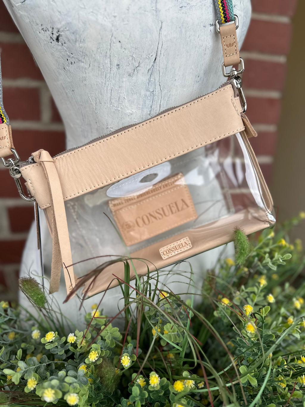A clear plastic crossbody bag with a genuine leather trim and woven strap, displayed on a white mannequin torso with green foliage and a brick wall in the background. The brand name 'CONSUELA' is visible on the leather tag and strap.