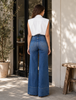 Woman wearing blue jeans and a white top standing in a sunlit outdoor setting.