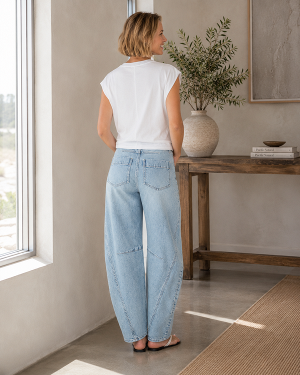 Woman wearing light blue jeans and a white top standing in a room with a wooden table and plant.