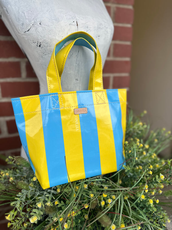 A glossy tote bag with bold vertical yellow and light blue stripes, displayed on a white mannequin bust surrounded by green foliage and small yellow flowers, with a brick wall in the background.