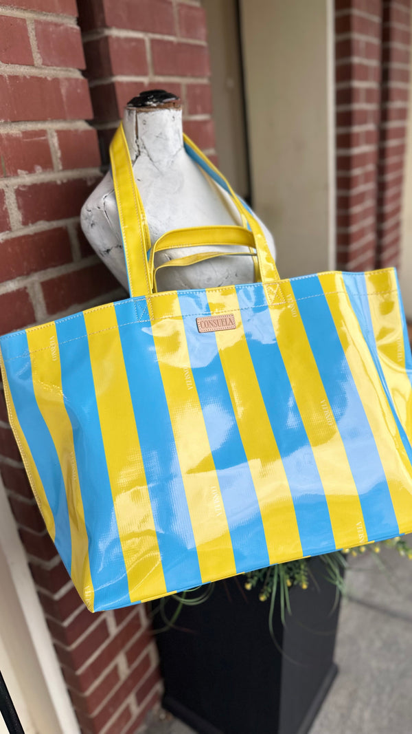A glossy vinyl tote bag with bold vertical yellow and light blue stripes, hanging from a white mannequin torso outdoors next to a red brick wall.