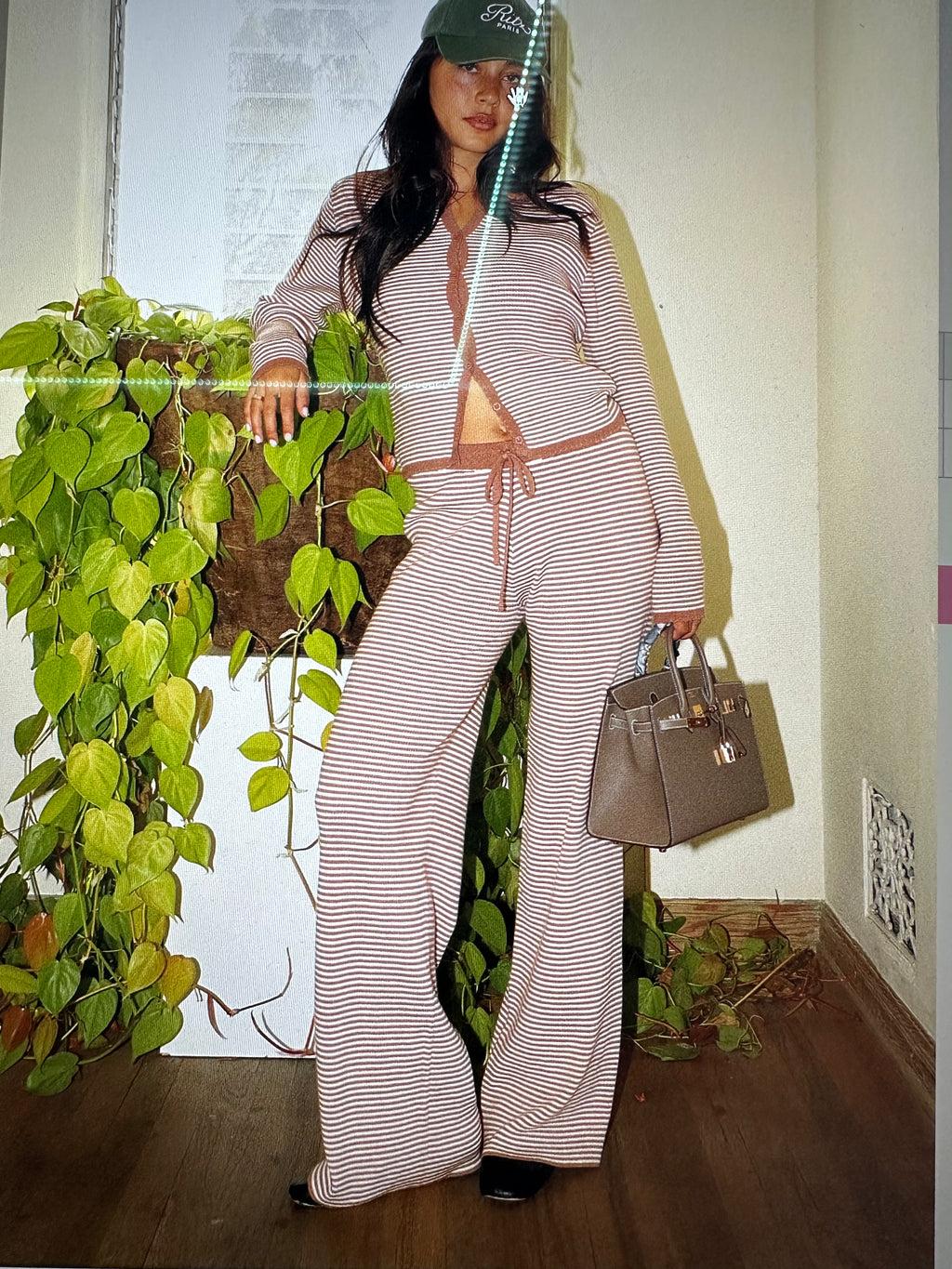 Woman in a striped outfit holding a handbag, standing next to a plant indoors.