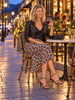 A woman sits at an outdoor cafe table at dusk, wearing a black leather moto jacket and a leopard-print midi skirt. She holds a glass of red wine, with warm streetlights and cafe ambiance in the background.