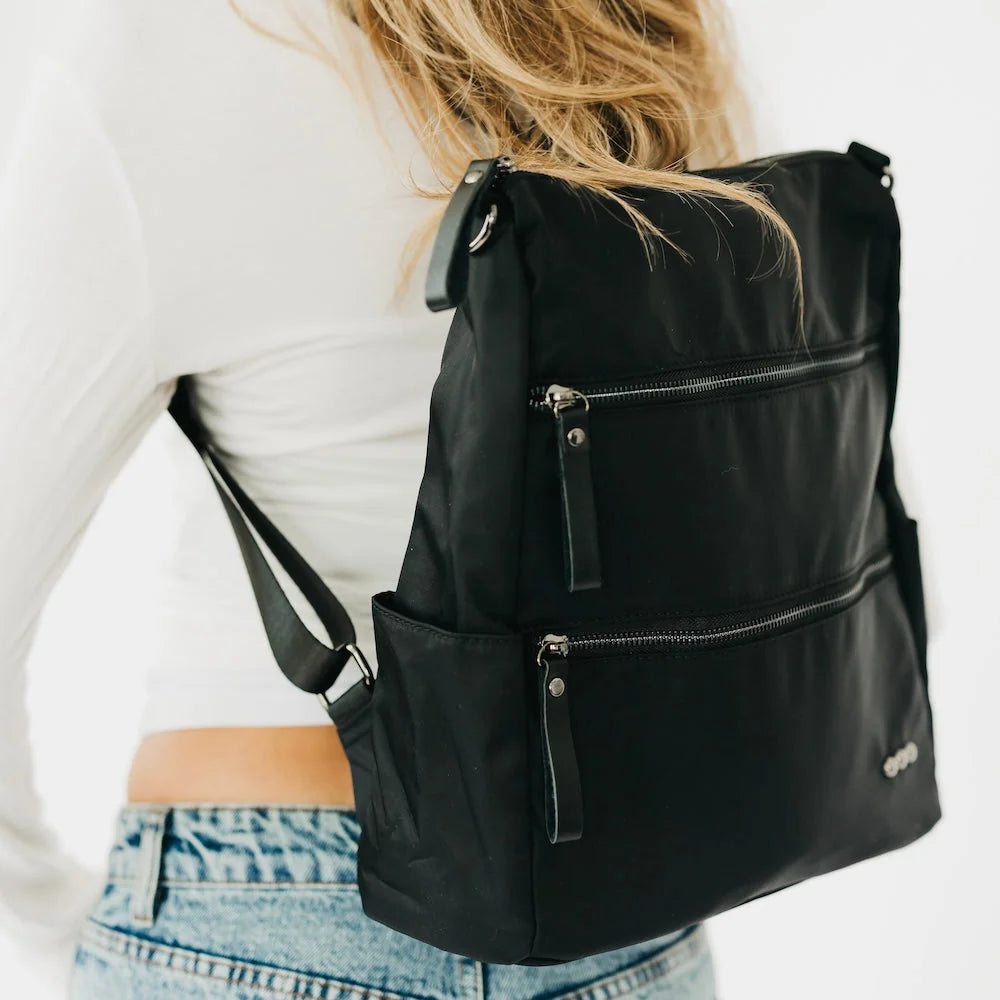 A black nylon shoulder bag with multiple zippered pockets, shown worn on the back of a person wearing a white long-sleeve top and light blue jeans.