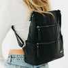 A black nylon shoulder bag with multiple zippered pockets, shown worn on the back of a person wearing a white long-sleeve top and light blue jeans.