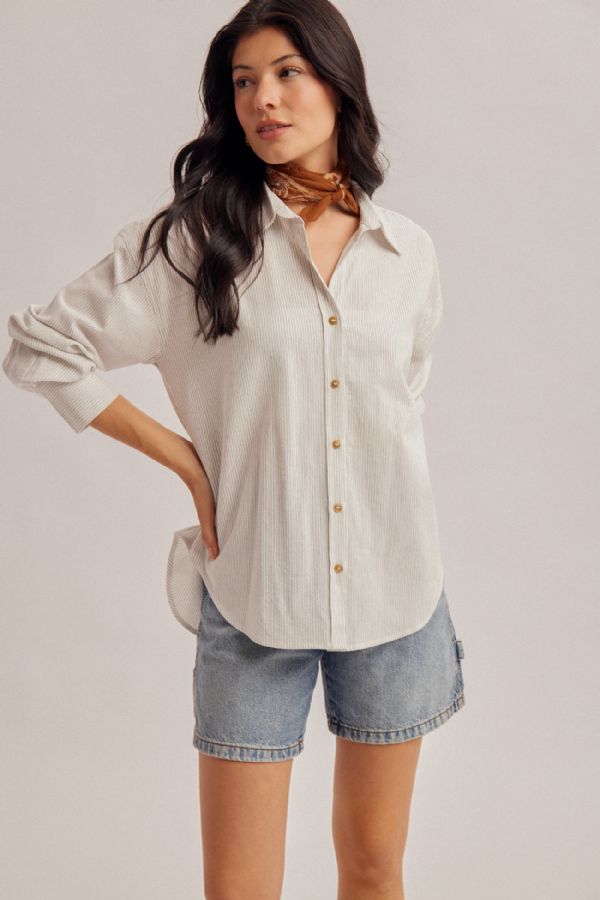 A woman modeling a pinstriped button-up shirt with light-wash denim shorts and a brown neck scarf, set against a plain light gray studio background.
