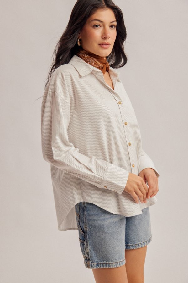A woman wearing a light beige long-sleeve button-up shirt with a subtle textured pattern, styled with denim shorts and a brown patterned neck scarf, against a solid light gray studio background.