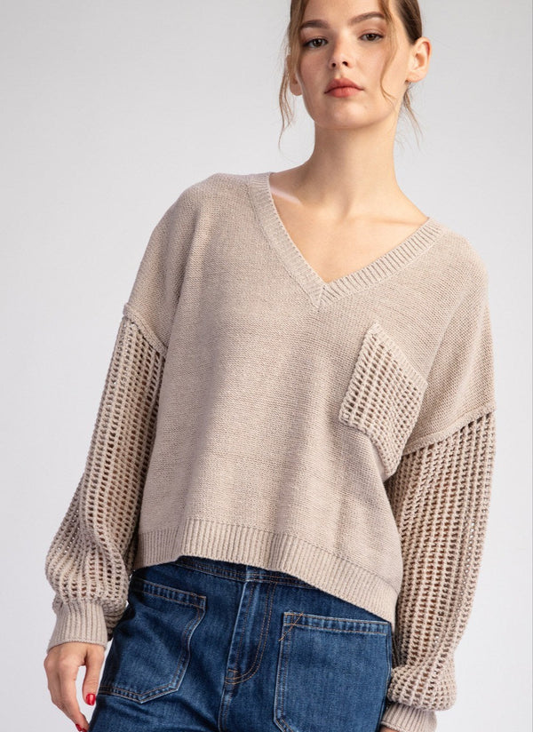 A female model wearing a long sleeve beige sweater with a ribbed v-neck and a front chest pocket, paired with blue jeans.