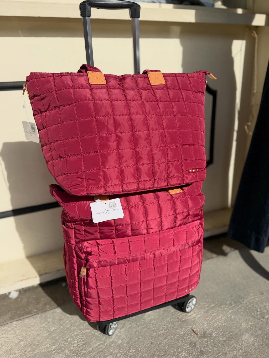 A red quilted rolling suitcase with a matching top tote bag, both featuring light brown leather accents and white price tags, set against a beige wall and concrete floor.