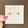 Gold flower earrings on a 'SALT' branded card held by a hand with pink nail polish.