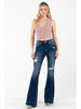A woman modeling a pair of dark blue high-waisted flare jeans with distressed details and ripped knees, paired with a dusty pink ribbed tank top, against a plain white studio background.